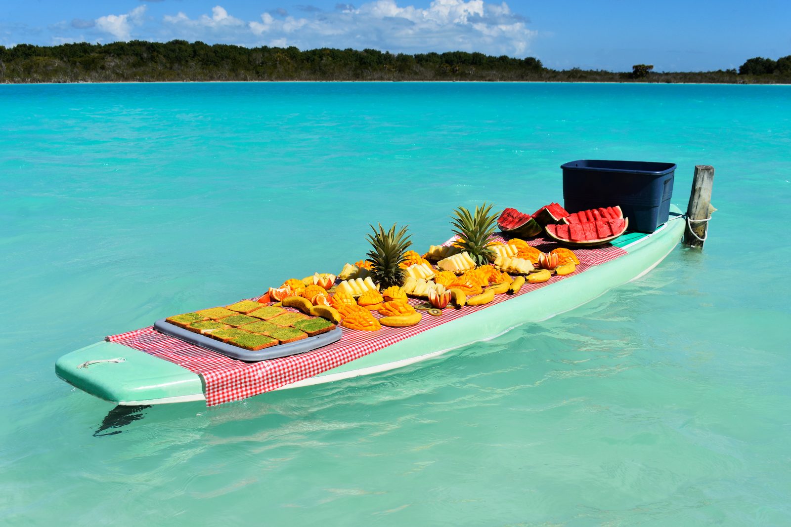 A floating paddleboard spread with sliced tropical fruit and pan de elote on the calm turquoise lagoon