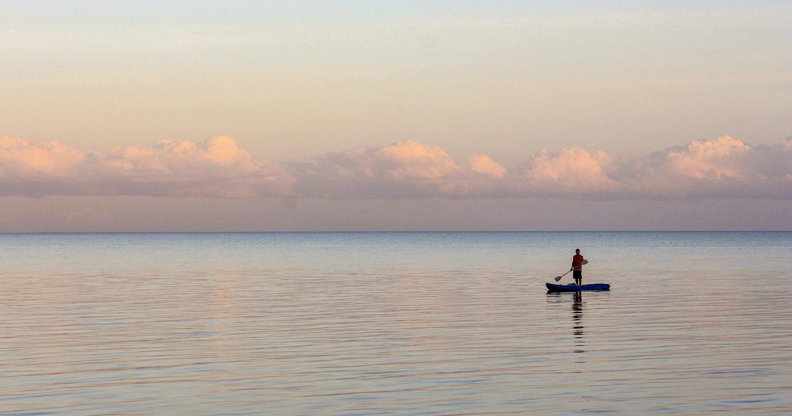 A lone paddleboarder gliding across glassy water at dawn under a pastel sky