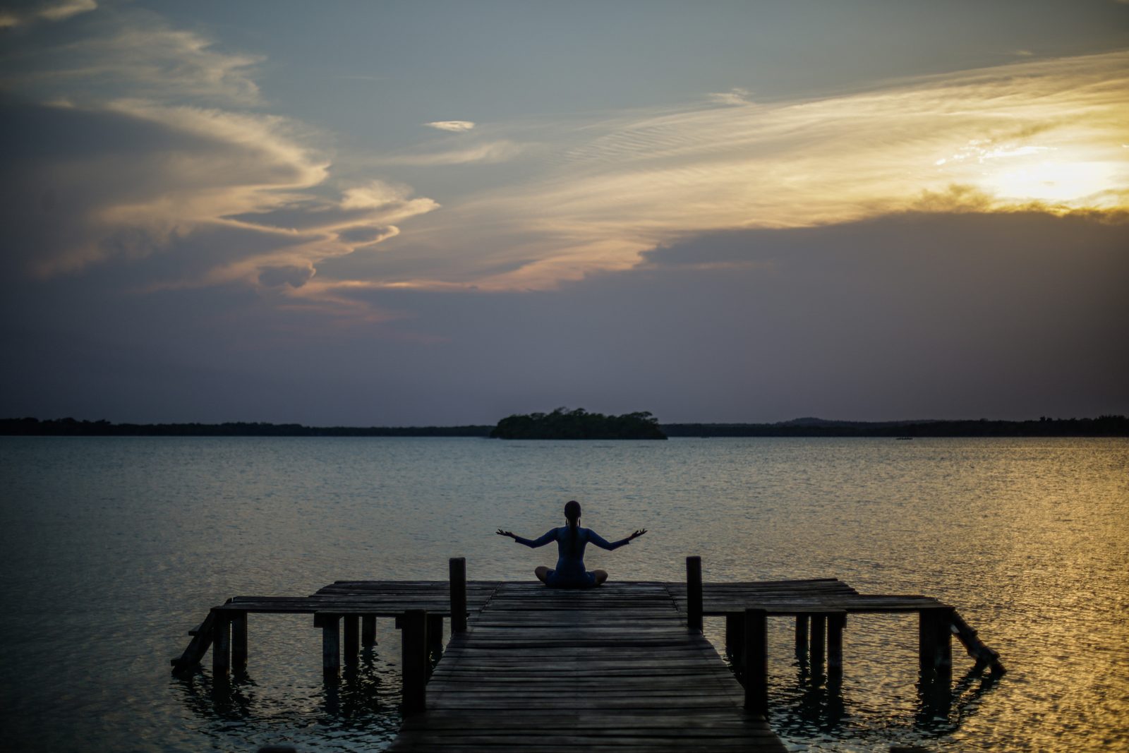 A person sitting in meditation at the end of a wooden dock as the sun sets over the lagoon