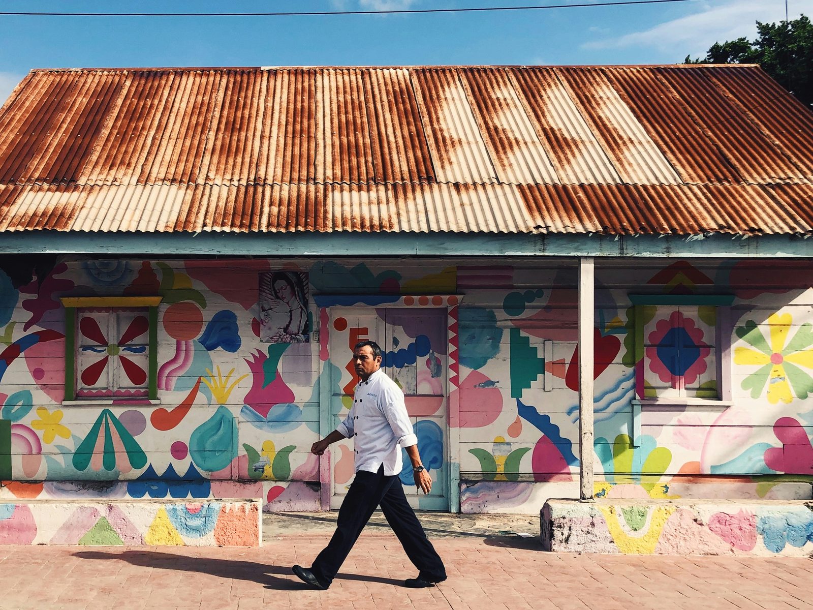 A chef walking past a clapboard house painted with bright folk-art murals under a rusted tin roof